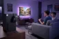 Preview: Stimmungsvolles Gaming-Zimmer Setup mit einem Spieler auf dem Sofa, einem gro&szlig;en TV und einer Xbox Series X, an der die schwarze anyprint3d RePlay Stand Controller-Halterung montiert ist.